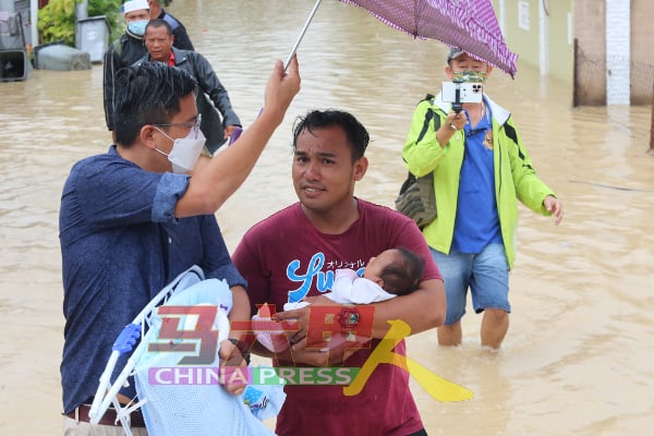 灾民抱着刚满月的婴儿涉水离开灾区，格西当州议员谢守钦（左）给予协助，帮忙拿东西及撑伞，避免淋湿孩子。