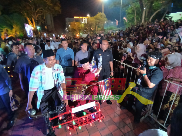 依斯迈沙比里（坐者）乘坐三轮车进入庆典会场期间，向观众席民众挥手示意。